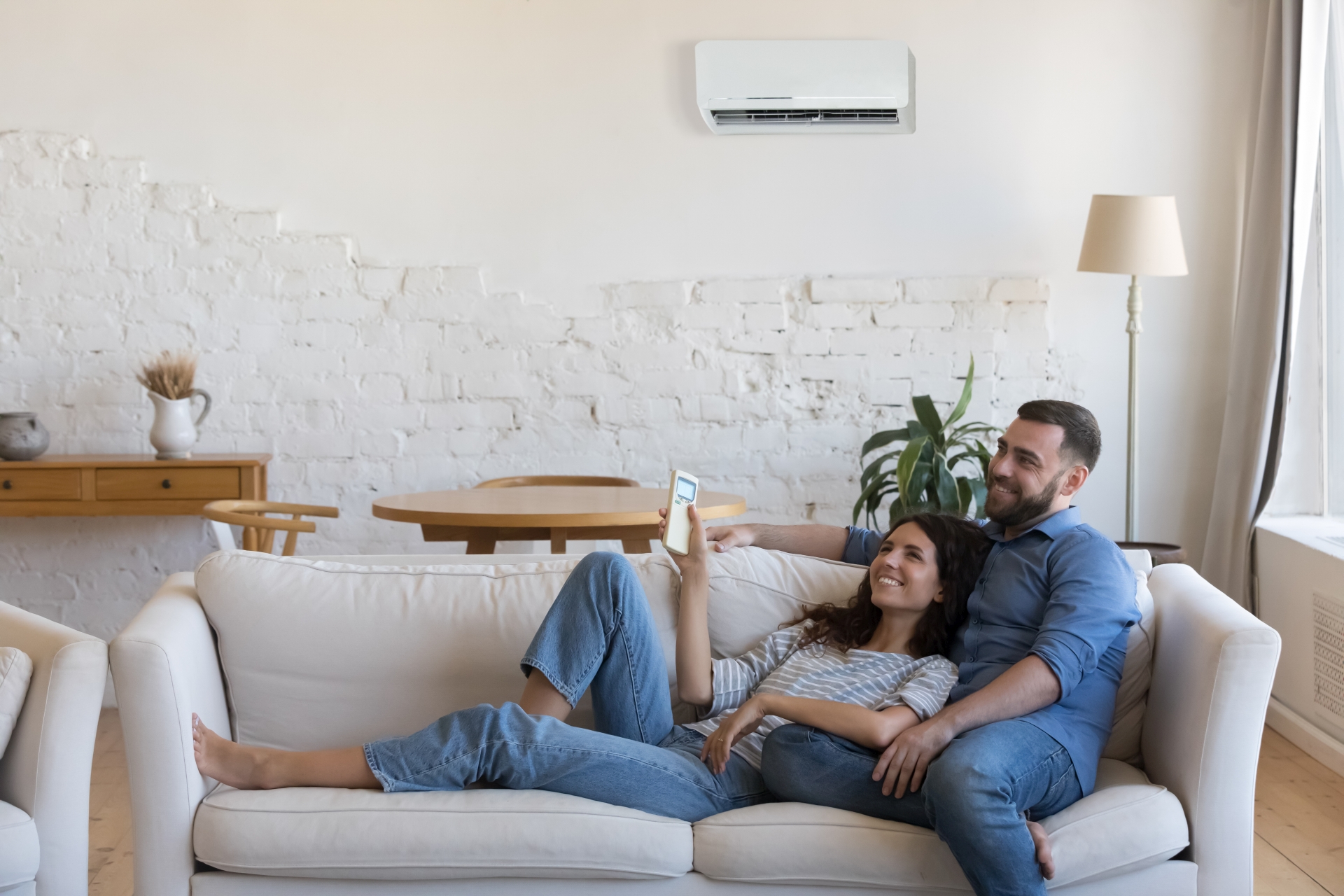 Couple enjoy fresh conditioned air inside cozy living room. Carefree wife holds remote controller adjust comfortable temperature relaxing on couch with husband at home. Tech for comfort life concept